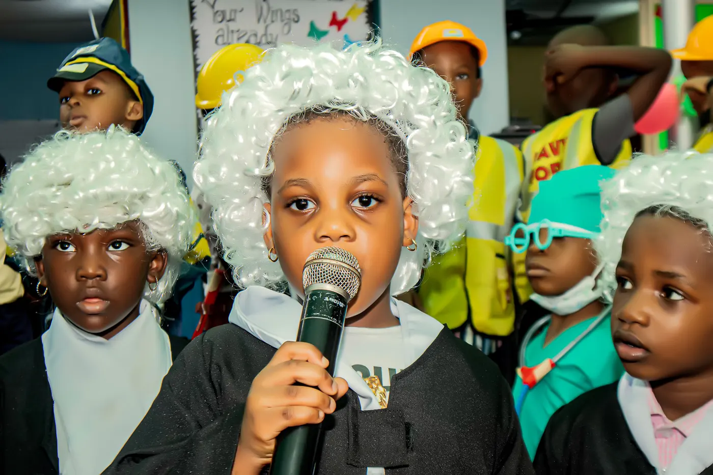A nursery pupil in a judge’s wig speaking into a microphone, surrounded by classmates in costume