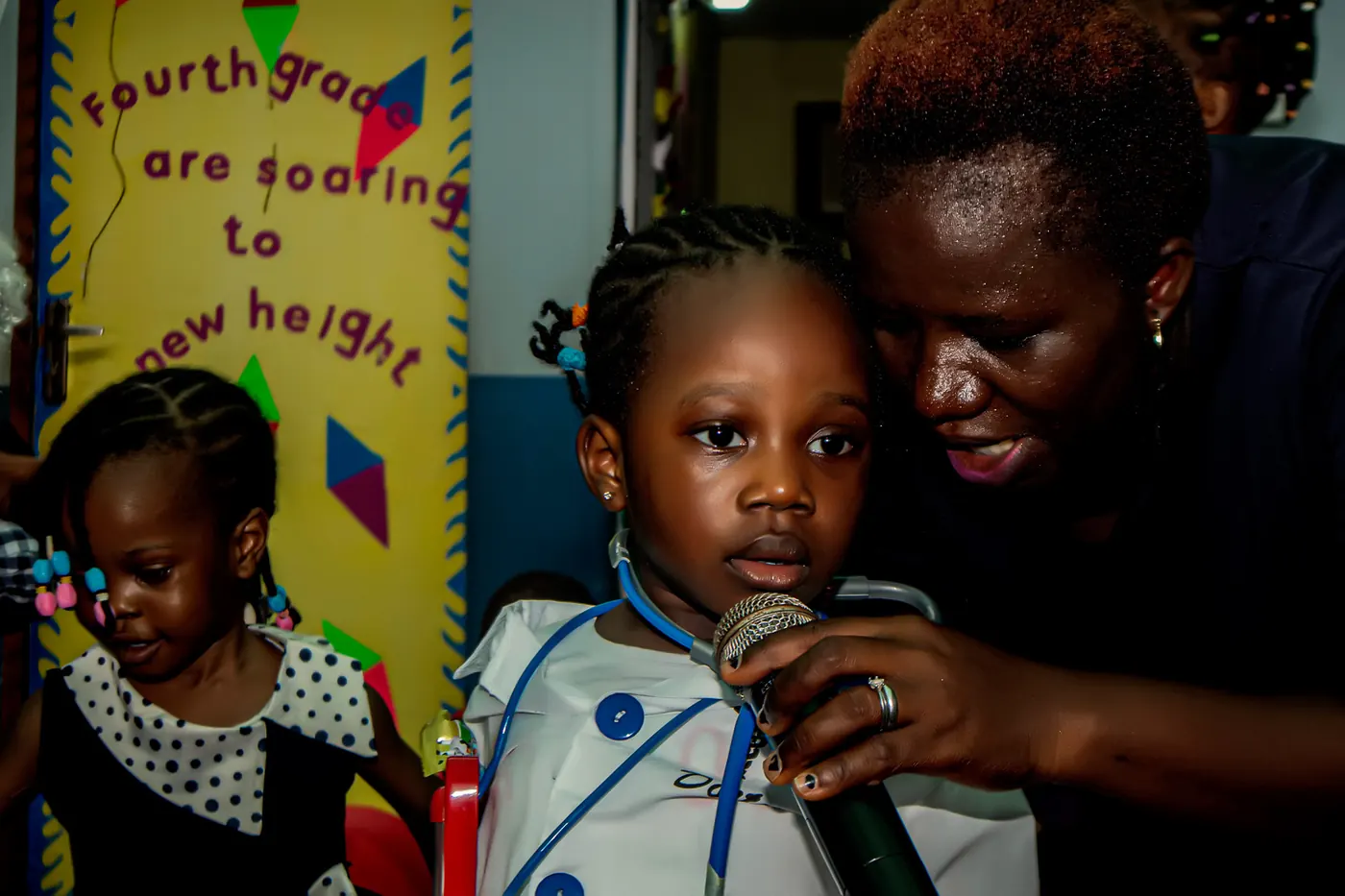 A young creche pupil speaking into a microphone with her teacher beside her
