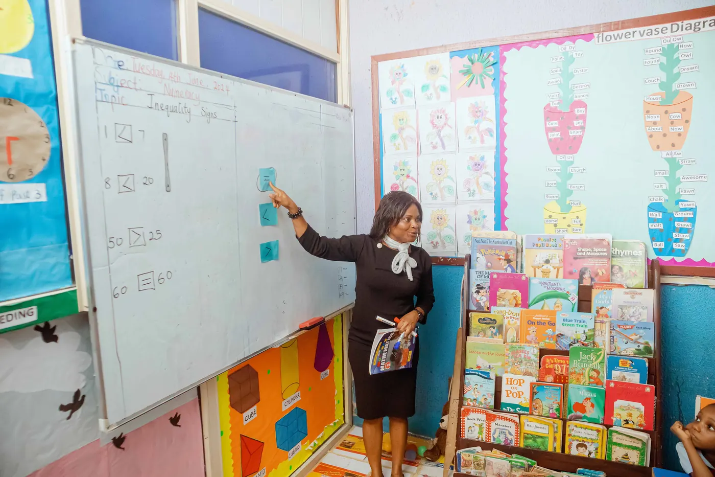 A teacher explaining a lesson at the whiteboard beside a shelf of picture books
